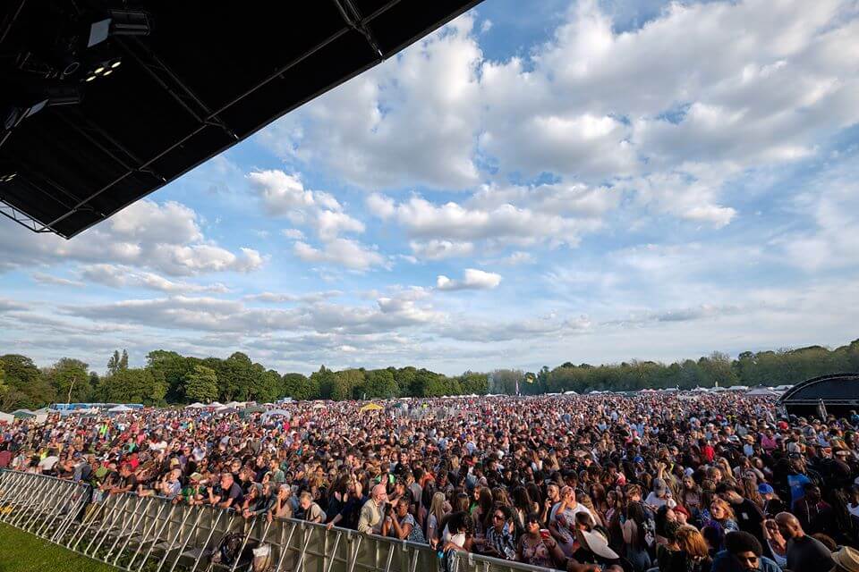 A packed crowd gathered in front of a stage in Sefton Park for the Africa Oyé festival. There are some large trees in the distance and clouds in the sky. 