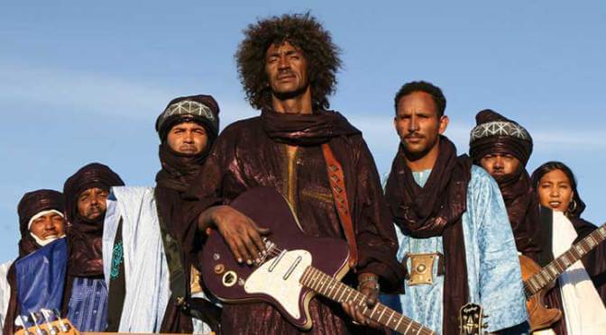 An image of a band standing outside, with a blue sky behind them. All seven members are looking towards the camera. Some are holding guitars. 